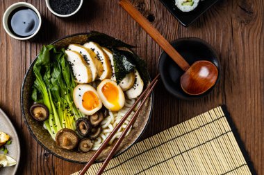 Geleneksel ramen ve domuz pastırması veya tavuk. Udon ya da ramen erişteli. Klasik kâselerde servis edilmiş. Arka planda Gyoza köftesi ve mantar var. Doğal ahşap zemin.