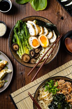 Geleneksel ramen ve domuz pastırması veya tavuk. Udon ya da ramen erişteli. Klasik kâselerde servis edilmiş. Arka planda Gyoza köftesi ve mantar var. Doğal ahşap zemin.