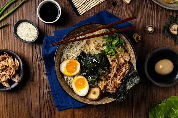 Geleneksel ramen ve domuz pastırması veya tavuk. Udon ya da ramen erişteli. Klasik kâselerde servis edilmiş. Arka planda Gyoza köftesi ve mantar var. Doğal ahşap zemin.