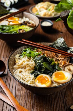 Geleneksel ramen ve domuz pastırması veya tavuk. Udon ya da ramen erişteli. Klasik kâselerde servis edilmiş. Arka planda Gyoza köftesi ve mantar var. Doğal ahşap zemin.