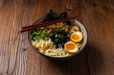 Geleneksel ramen ve domuz pastırması veya tavuk. Udon ya da ramen erişteli. Klasik kâselerde servis edilmiş. Arka planda Gyoza köftesi ve mantar var. Doğal ahşap zemin.