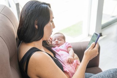 Akıllı telefonuyla çalışan genç bir kadın bebeğiyle kollarında uyuyor.