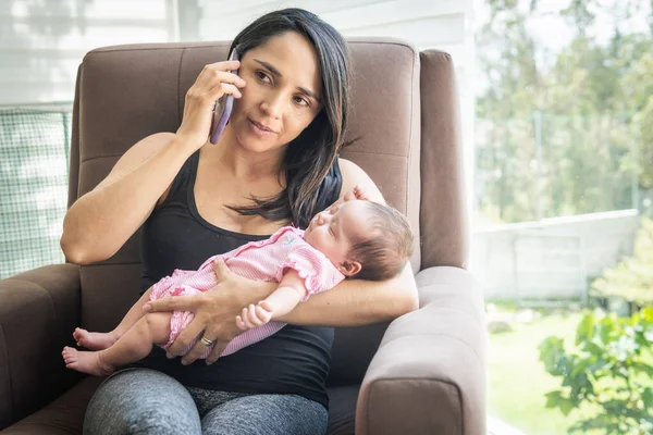 Latin mother using talking on cell phone with her sleeping baby in her arms at home