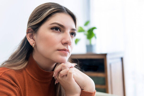 close-up of young woman with thoughtful look and her hands on her jaw and staring in the living room of her house