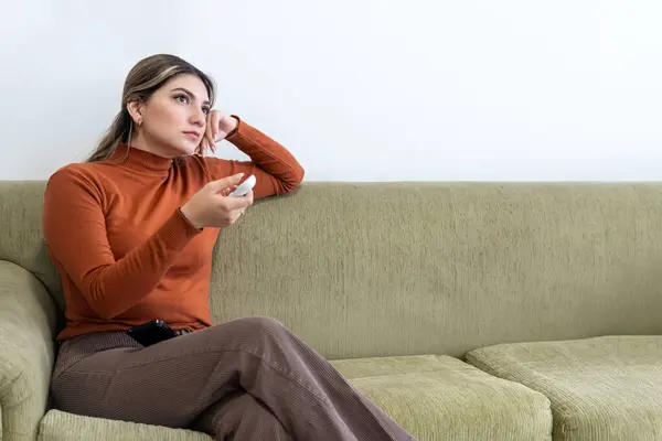 stock image Young Latina woman in her 20s sitting on a couch pointing a remote control at an electronic device