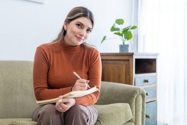 Young female psychologist looking at camera with her notebook and a pen in her office clipart