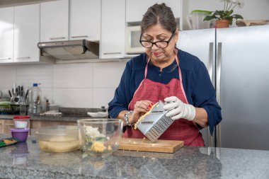 Kıdemli Latin kadın rendelenmiş yeşil muz rendeliyor. O bir önlük ve eldiven giyiyor. Mutfakta bir buzdolabı, mikrodalga fırın ve saksı bitkisi var.