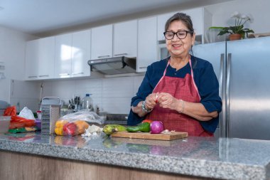 Mutfakta soğan, biber ve domates gibi sebzelerin yanında sarımsak soyan son sınıf Latin bir kadın. Kadın yemek yaparken gülümsüyor.