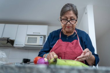 Bir kadın mutfakta sebze doğruyor. Üzerinde mavi bir önlük ve kırmızı bir bandana var.