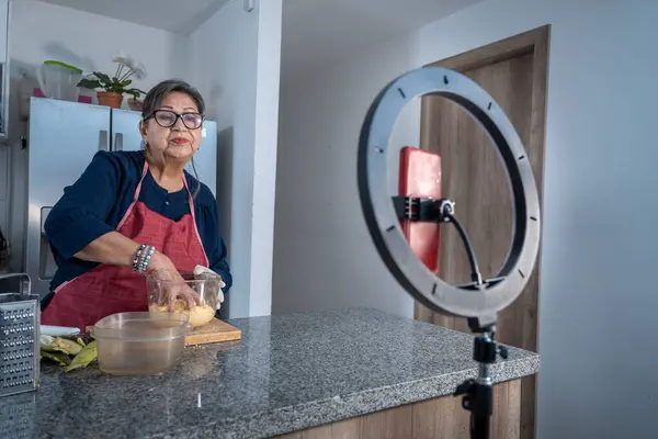 Üst düzey Latin kadın sosyal medya için ışık ve cep telefonuyla yemek kursunu videoya kaydediyor. Modern yetişkin kadın dijital içerik üretiyor