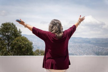 Sırtı dönük bir kadın, elinde cep telefonuyla bir dağın önünde kollarını açıyor. Sabahları başarılı ve liderlik pozisyonunda.