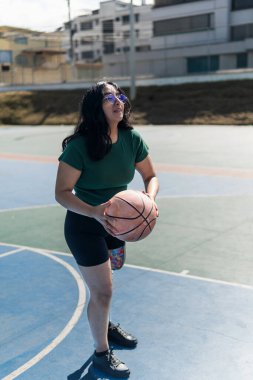 Protez bacaklı atletik kadın dışarıda basketbol oynuyor, topu tutuyor ve yukarı bakıyor.