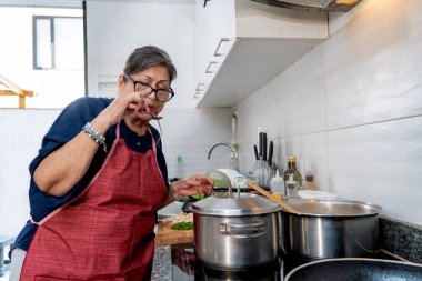 Yaşlı İspanyol kadın önlük giyiyor modern mutfakta kaşıkla yemek tadıyor.