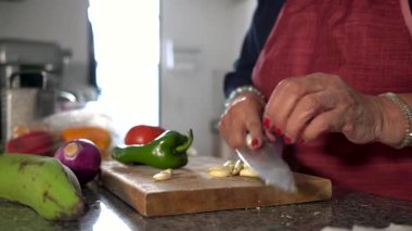 Yaşlı bir kadın, bir mutfak ortamında taze sebzelerle çevrili ahşap kesme tahtasına sarımsak ustalıkla doğruyor.