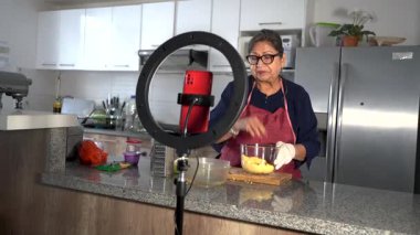 Önlük ve eldiven giyen yaşlı bir kadın ev mutfağında malzeme hazırlıyor, akıllı telefon ve halka ışığıyla video dersi kaydediyor.