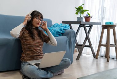 Kulaklıkla müzik dinleyen ve laptopla çalışan bir kız oturma odasında yerde oturuyor.