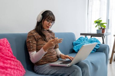 Latin bir kadın, kulaklıkla müzik dinlerken kredi kartıyla ödeme yapıyor ve evdeki kanepede laptopuyla online alışveriş yapıyor.
