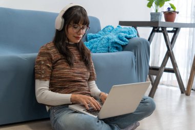 Laptop ve kulaklık kullanarak uzaktan çalışan, evinin keyfini çıkaran oturma odasının zemininde oturan genç bir kadın.
