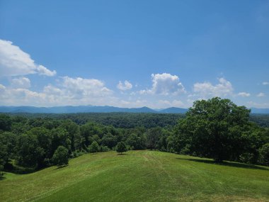 Bereketli yeşil bir manzaraya sahip yuvarlanan tepeler ve açık mavi gökyüzünün altındaki uzak dağlar dağınık bulutlar..