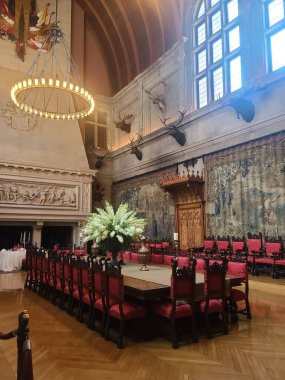 A grand dining hall featuring a long wooden table surrounded by ornate chairs. The room is adorned with large tapestries, mounted animal heads on the walls, and a large chandelier overhead. A floral arrangement is placed at the center of the table, a clipart