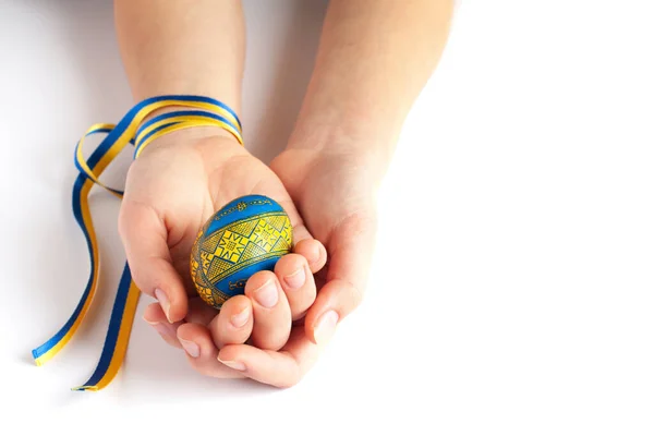 a blue and yellow Easter egg in the hands of a child with a patriotic ribbon on a white background