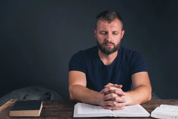 A man prays with folded hands. An open Holy Bible on the table. A young man with a beard.