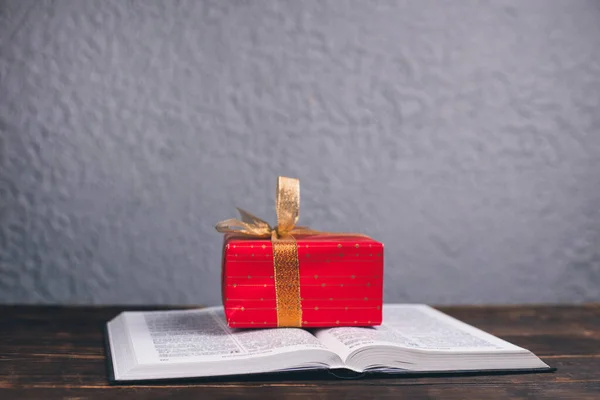 An open book of the Bible on the table. A gift on the Bible in a red box.