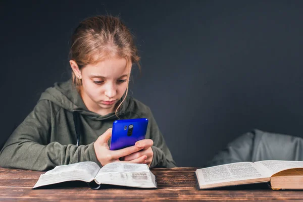 An open Bible on the table. A girl with a phone in her hands. Ignores books and plays video games on phone.