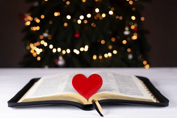 An open Bible on the table. The heart is a symbol of God\'s love for people. Prayer. On the background of the Christmas tree