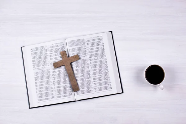 An open Holy Bible on the table. Wooden cross. Crucifix. A cup of coffee on the table.