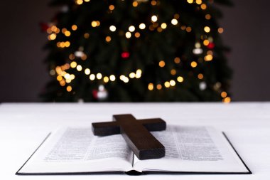 An open Bible on the table. Prayer. Wooden cross. Crucifix. Holy Scripture is the heart. On the background of the Christmas tree. Light and garlands