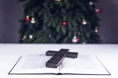 An open Bible on the table. Prayer. Wooden cross. Crucifix. Holy Scripture is the heart. On the background of the Christmas tree. Light and garlands