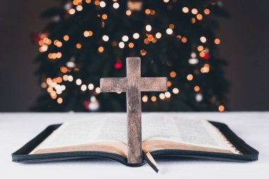 An open Bible on the table. Prayer. Wooden cross. Crucifix. Holy Scripture is the heart. On the background of the Christmas tree. Light and garlands