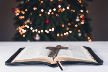 An open Bible on the table. Prayer. Wooden cross. Crucifix. Holy Scripture is the heart. On the background of the Christmas tree. Light and garlands