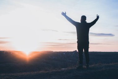 Elleri havada bir adam dua ediyor. Günbatımı gökyüzünün arka planında. Günahlar için tövbe. Diz çöküp Tanrı 'ya dua etmek. İbadet ve övgü.