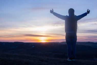 Ellerini kaldırmış bir kız dua ediyor. Günbatımı gökyüzünün arka planında. Günahlar için tövbe. Diz çöküp Tanrı 'ya dua etmek. İbadet ve övgü.