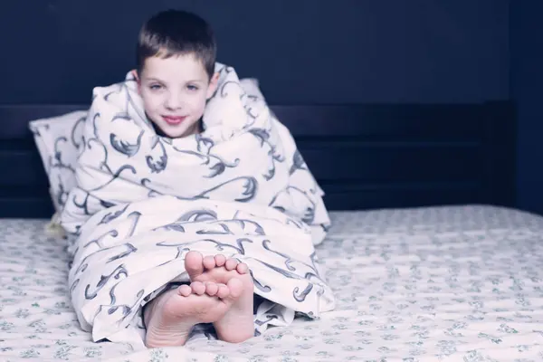 Bare feet of a child. A child in pajamas is smiling. The child is wrapped in a blanket. The boy is sleeping in bed. Foot and leg