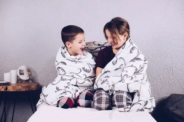 Two children wrapped in a blanket on the bed. Happy brother and sister hugged. Children smiling in pajamas.