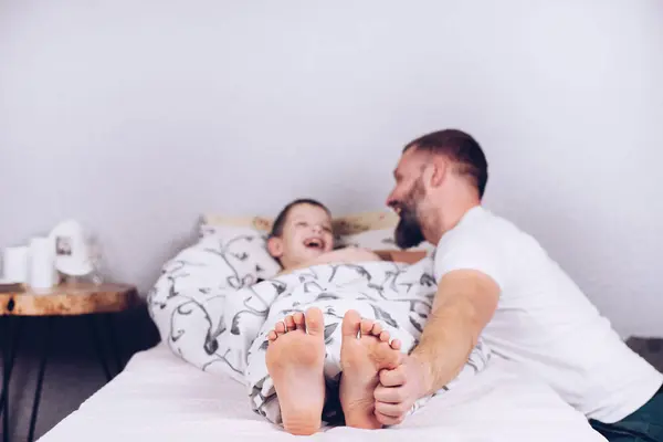 A child with bare feet is lying in bed. Dad tickles his son. Tickling the heels. Cheerful and joyful laughter of a happy child.