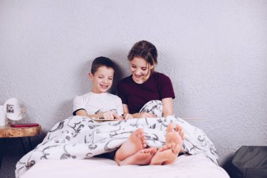 Erkek ve kız kardeş birlikte bir kitap okudular. Yatakta çıplak ayakla mutlu ve neşeli çocuklar. Bacakları bir battaniye ile kaplıdır. Çarşaflar.