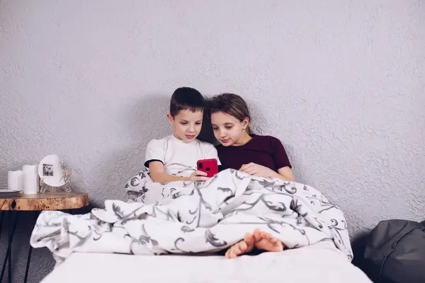 Two teenagers are playing on the phone. Addiction to the phone. Brother and sister playing video games. Children under the blanket. Lying on the bed.