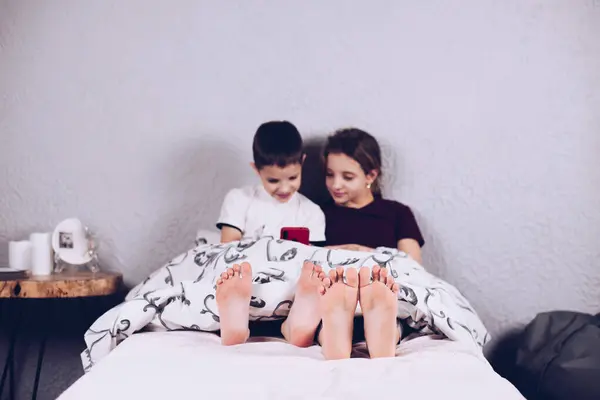 Two teenagers are playing on the phone. Addiction to the phone. Brother and sister playing video games. Children under the blanket. Lying on the bed.