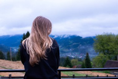 Saçları dökülmüş genç bir kız yüzünü kapatıyor. Dağların arka planında.