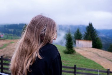 Saçları dökülmüş genç bir kız yüzünü kapatıyor. Dağların arka planında.