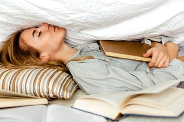 Beyaz tişörtlü tatlı sarışın kadın yatak odasındaki yatakta kitap okuyor. Örtünün altında bir kitapla mankenlik yap. Önplanda birçok kitap
