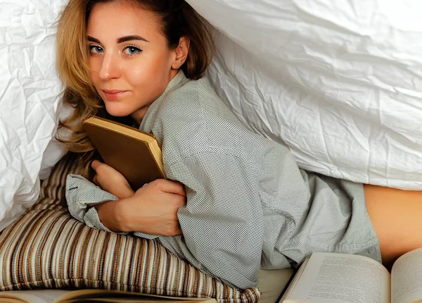漂亮的金发女人穿着白衬衫躺在卧室的床上 模型下的毛毯覆盖一本书 前景中的书很多 — 图库照片