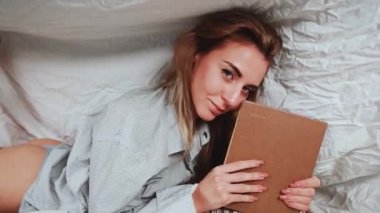 Beyaz tişörtlü tatlı sarışın kadın yatak odasındaki yatakta kitap okuyor. Örtünün altında bir kitapla mankenlik yap. Önplanda birçok kitap