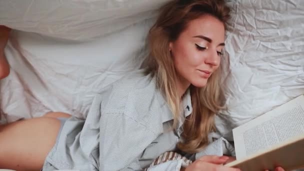 漂亮的金发女人穿着白衬衫躺在卧室的床上 模型下的毛毯覆盖一本书 前景中的书很多 — 图库视频影像