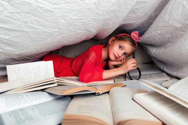 Cute 6-7 years old girl reads a book. Little girl at home on the bed in red dress under a blanket cover with a many books on foreground. Search information in book with magnifying glass