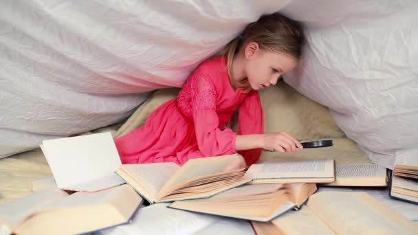可爱的6 7岁的女孩读一本书 家里的小女孩穿着红衣服躺在床上 盖着毛毯 前面放着许多书 用放大镜搜索书籍中的信息 — 图库视频影像